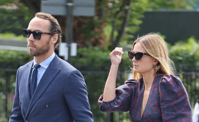 A couple in formal attire and sunglasses walking outdoors, with greenery in the background.