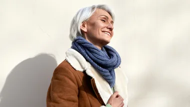 Woman smiling, wearing a brown coat and blue scarf, standing against a light wall in winter sunlight.