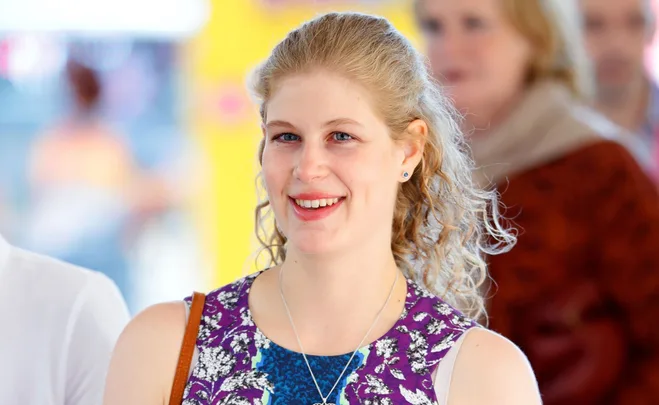A young woman with curly blonde hair smiles, wearing a colorful patterned dress and a necklace, with a blurred background.