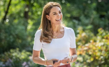 A woman in a white top smiling outdoors with trees in the background.