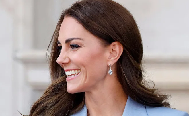 Woman with long brown hair smiling, wearing elegant earrings and a blue coat.
