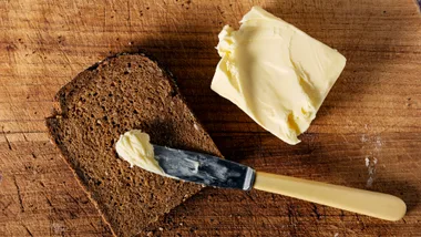 Slice of bread with a knife spreading butter on a wooden board.
