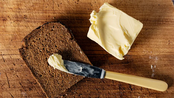Slice of bread with a knife spreading butter on a wooden board.