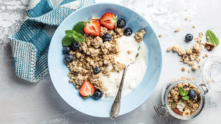Bowl of yogurt topped with granola, strawberries, blueberries, and mint with a spoon, next to a jar of mixed granola.