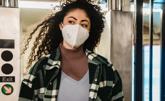 Person with curly hair wearing a face mask and plaid shirt in a public setting near an exit sign.