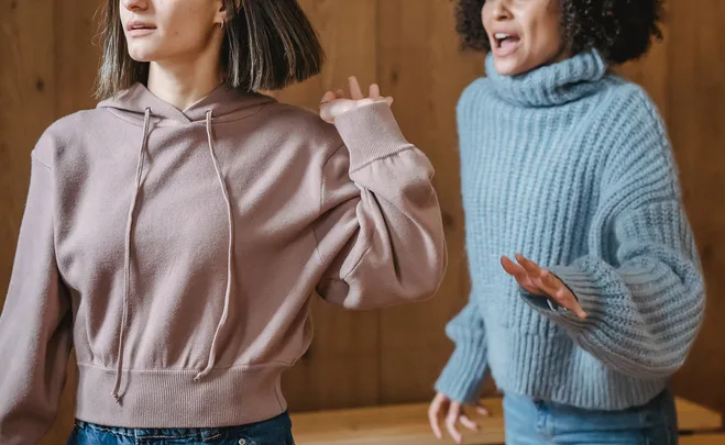 Two women in sweaters arguing, one gesturing with frustration.