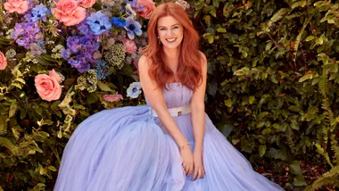 Woman in a flowing lavender dress sits smiling in front of lush greenery and colorful flowers.