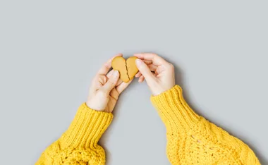 Hands in yellow sweater holding a broken heart-shaped cookie against a gray background.