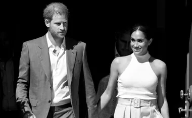 Prince Harry and Meghan Markle smiling and holding hands, exiting a building.