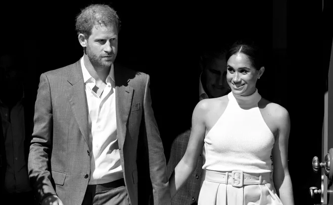 Prince Harry and Meghan Markle smiling and holding hands, exiting a building.