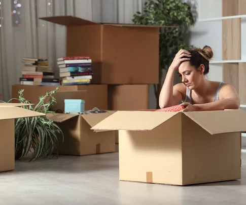 Woman looking stressed while packing boxes in a room, surrounded by books and plants.
