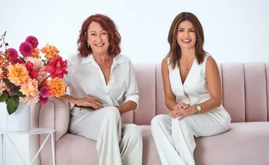 Two women in white outfits sitting on a pink sofa, smiling, next to a bouquet of colorful flowers.