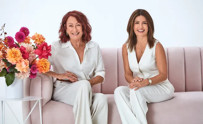 Two women in white outfits sitting on a pink sofa, smiling, next to a bouquet of colorful flowers.