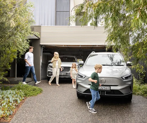 Family in driveway with electric cars; parents and kids walking near a silver SUV and a white car in front of garage.