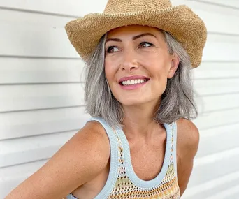 Woman in a straw hat smiling, wearing a sleeveless crochet top, stands in front of a light-colored wall.