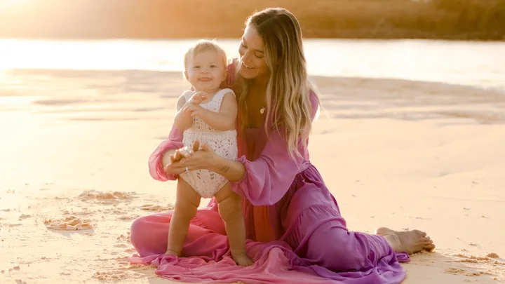 A woman in a flowing pink dress sits on a sandy beach, holding a smiling baby in a white outfit in warm sunlight.