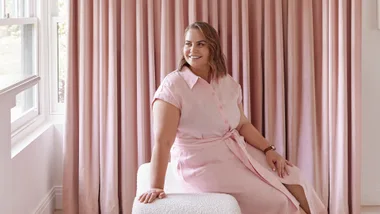 Woman in pink dress sitting on a white chair, smiling, with pink curtains in the background.
