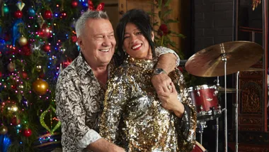 A smiling couple in festive attire poses in front of a decorated Christmas tree and musical instruments.