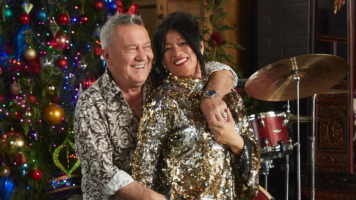 A smiling couple in festive attire poses in front of a decorated Christmas tree and musical instruments.