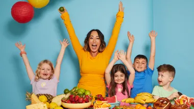A woman in yellow and five kids joyfully raise hands, surrounded by fruits and colorful lanterns on a blue background.