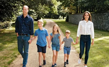 A smiling family of five walking on a tree-lined path in casual outfits on a sunny day.
