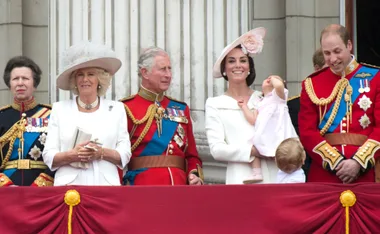 Royal family on balcony in formal attire during an event, with a child looking up in the foreground.
