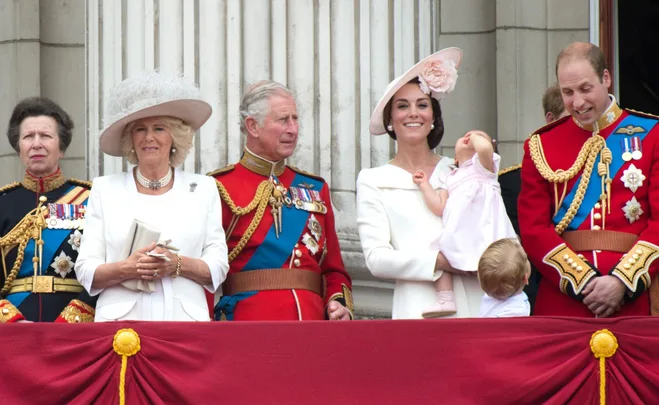 Royal family on balcony in formal attire during an event, with a child looking up in the foreground.