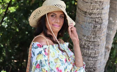 A woman in a floral dress and straw hat stands by a tree, smiling gently with greenery in the background.