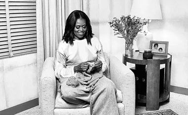 Michelle Obama sits knitting in a cozy chair, with flowers and a framed photo on a side table.