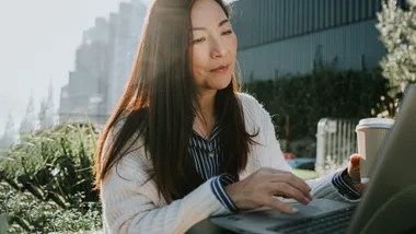 Woman working on a laptop outdoors, holding a coffee cup, with sunlight and plants in the background.