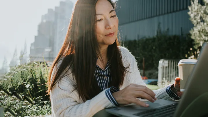 Woman working on a laptop outdoors, holding a coffee cup, with sunlight and plants in the background.