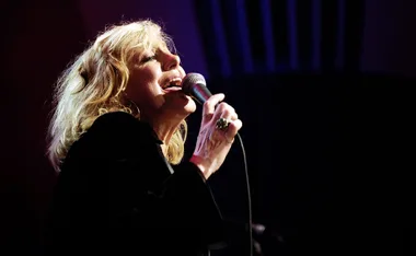 Singer passionately performing on stage with microphone, dressed in black, illuminated by stage lights.