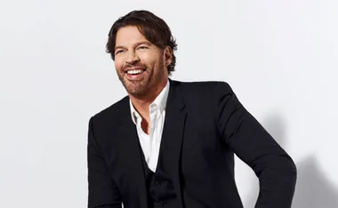 A man in a black suit with a white shirt, smiling and looking to the side against a light background.