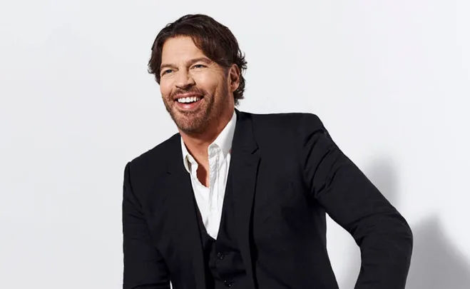 A man in a black suit with a white shirt, smiling and looking to the side against a light background.