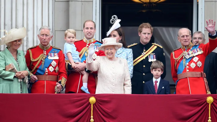 A group of royals standing on a balcony, dressed formally, with one woman waving to the crowd below.