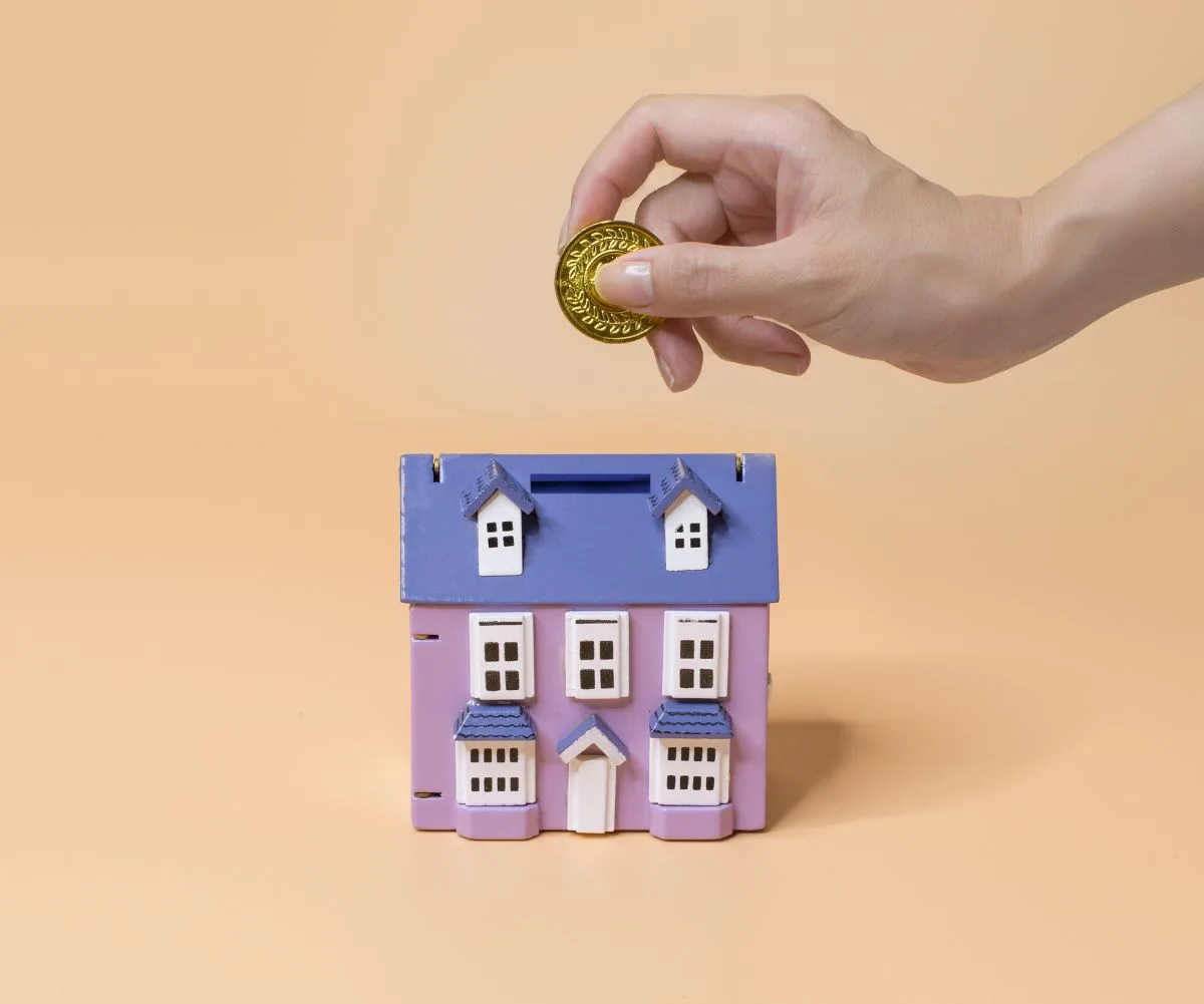 Hand placing a coin into a purple house-shaped piggy bank on a beige background.