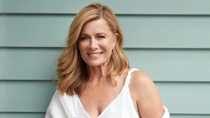 A woman with blonde hair smiles against a light blue wall, wearing a white sleeveless top and a white cardigan.
