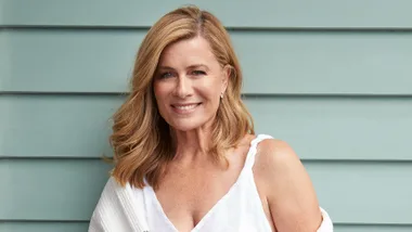 A woman with blonde hair smiles against a light blue wall, wearing a white sleeveless top and a white cardigan.