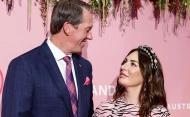 A man in a blue suit and pink tie smiles at a woman in a striped dress with a beaded headband, against a pink flowering backdrop.