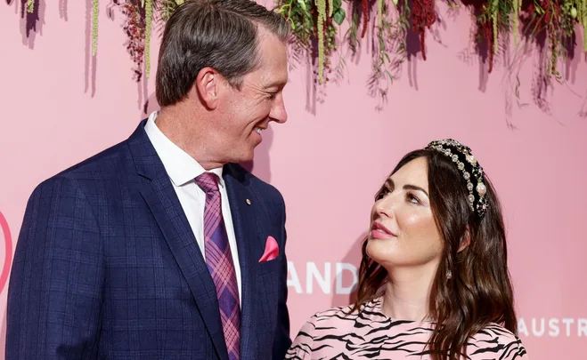 A man in a blue suit and pink tie smiles at a woman in a striped dress with a beaded headband, against a pink flowering backdrop.