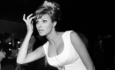 Black and white image of a glamorous woman in a white dress adjusting her hair, wearing a tiara, with people in the background.
