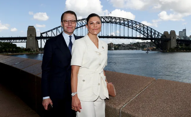 Two people in formal attire stand in front of the Sydney Harbour Bridge on a sunny day.