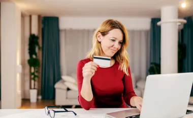 Woman in red using laptop, holding credit card, in a bright room with blue curtains.