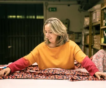 A woman in an orange sweater works with colorful fabrics in a workshop, surrounded by shelves and materials.