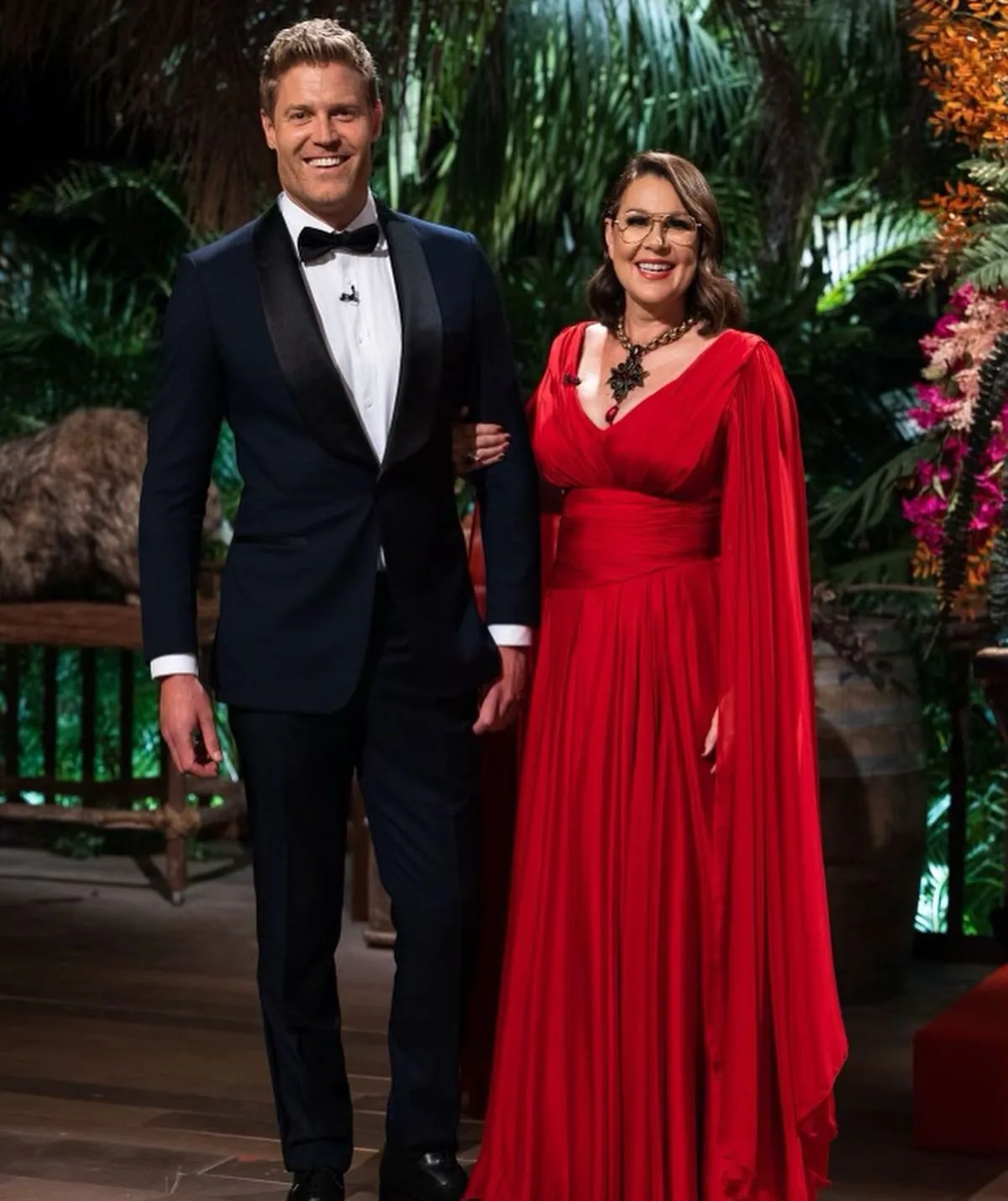 A man in a tuxedo and a woman in a red gown, smiling, standing together on a lush, vibrant set.
