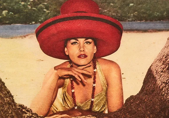 Woman in a large red hat and yellow swimsuit lying by a tree on the beach.