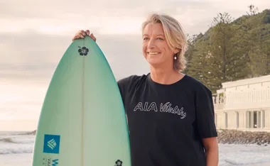 A woman with short blonde hair smiles, holding a turquoise surfboard with a hibiscus design, at the beach during sunset.