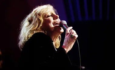Singer performing passionately on stage with a microphone against a dark background.