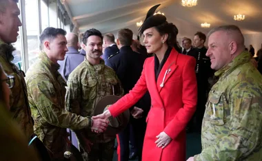 A woman in a red coat and black hat shakes hands with soldiers in camouflage uniforms at an indoor event.