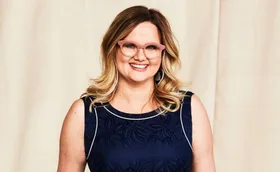 A woman wearing pink glasses and a navy dress smiles against a beige background.